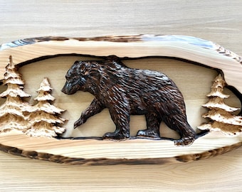 May include: A carved wooden wall hanging featuring a brown bear walking through a forest scene with two small pine trees. The bear is carved in a realistic style and the wood has a natural, rustic finish.