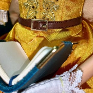 May include: A yellow dress with a brown belt and a gold embroidered design. The dress is being held by a person who is also holding a blue and gold book.