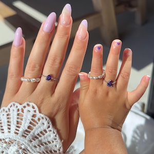 May include: Two hands, one adult and one child, each adorned with rings. The adult's hand features a pearl ring and a silver ring with a teardrop-shaped purple gemstone. The child's hand also has a pearl ring and a similar gemstone ring.