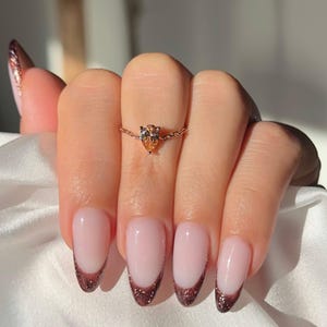 May include: A rose gold ring with a pear-shaped, light orange gemstone. The ring is on a chain. The nails are painted with a pink base and a dark glittery brown French tip.