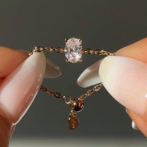 May include: A delicate gold chain ring featuring a sparkling oval-cut diamond. The ring's design includes a chain band and a small charm. The ring is held between two fingers.