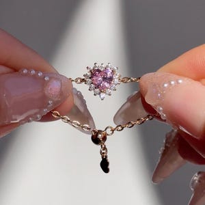 May include: A delicate gold-toned chain ring featuring a pink heart-shaped gemstone surrounded by smaller white gemstones. The ring's design includes a chain that adjusts for size, and a small charm.