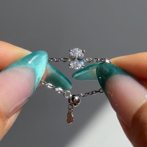 May include: A silver ring with an oval-shaped, clear gemstone. The ring's band is a delicate chain, and the gemstone is held in place by four prongs. The ring is being held up by a person's fingers with teal nail polish.