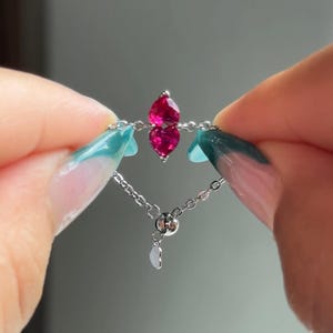 May include: A silver adjustable ring featuring two vibrant, pear-shaped, ruby-red gemstones. The ring's delicate chain design includes a small, heart-shaped charm. The gemstones are set close together, creating a striking visual contrast against the silver.