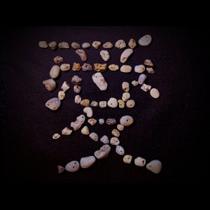 Könnte beinhalten: Ein japanisches Schriftzeichen aus kleinen, hellen Steinen vor schwarzem Hintergrund. Die Steine, in verschiedenen Formen und Größen, bilden das Symbol. Einige Steine haben Löcher. Das Bild hat eine minimalistische Ästhetik.