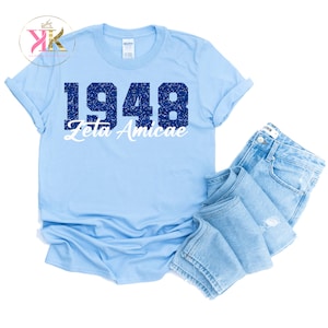 May include: A light blue t-shirt with the text "1948 Zeta Amicae" in a distressed navy blue and white font. The shirt is paired with folded light blue denim jeans. The t-shirt is a Gildan brand.