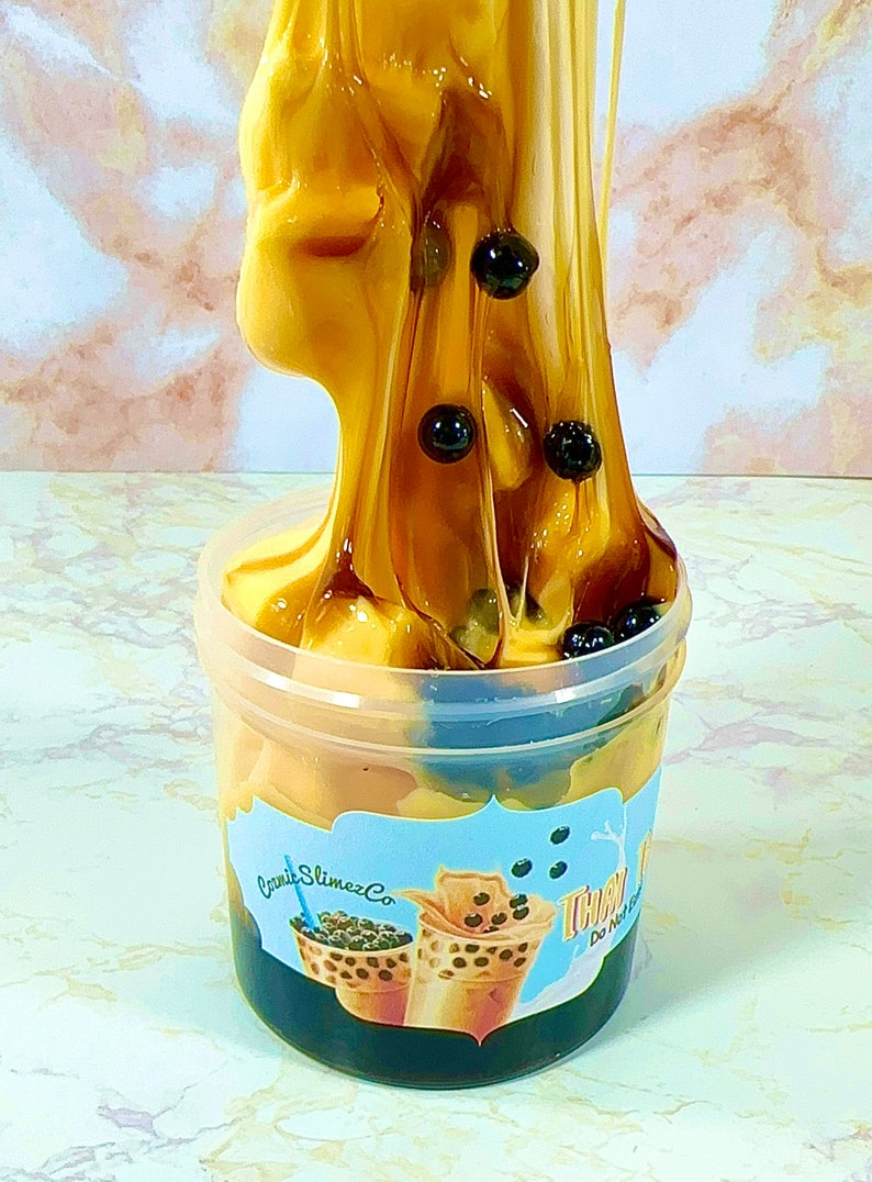 May include: A clear plastic container filled with brown and black slime with black boba pearls. The slime is being poured from the top of the container. The container has a blue and white label that says "Candi Slimez Co." and "Thai Tea Slime".