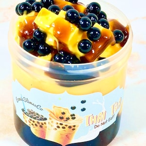 May include: A clear plastic container filled with a yellow and brown slime topped with black boba pearls. The container has a label that says "Comic SlimezCo Thai Tea Do Not Eat".