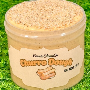 Peut inclure: Un pot en plastique transparent rempli d'une substance gluante de couleur beige, surmonté d'une couche de petits granulés brun clair. Le pot porte une étiquette avec le texte "Cozmic SlimezCo Churro Dough" et une illustration de churros. Le texte "DO NOT EAT!" est également présent.