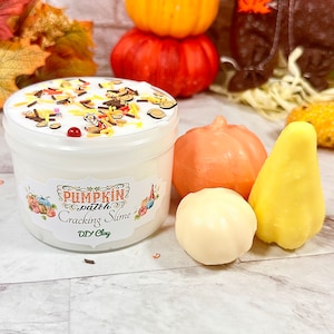 May include: A jar of white Pumpkin Patch Cracking Slime with colorful sprinkles and a decorative label. Beside the jar are small pumpkin and pear-shaped decorations. A stack of pumpkins in orange and yellow are in the background.