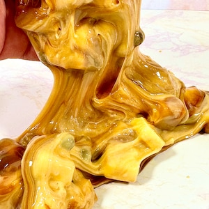 May include: Close-up of a brown and yellow slime with a swirled pattern. The slime is being stretched and pulled, revealing its gooey texture.
