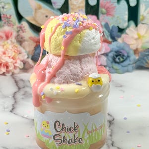 May include: A jar of "Chick Shake" with a layered dessert design. The top features yellow and pink scoops with pink drizzle and colourful sprinkles. A small chick-shaped decoration sits on the side. The jar has a label with a chick illustration.