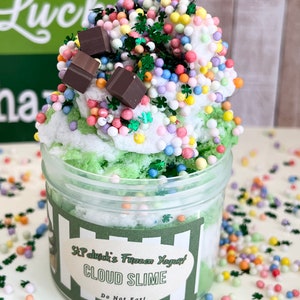 May include: A clear jar filled with green and white cloud slime topped with colorful sprinkles and chocolate pieces. The jar has a label that reads "St. Patrick's Frozen Yogurt Cloud Slime Do Not Eat!"
