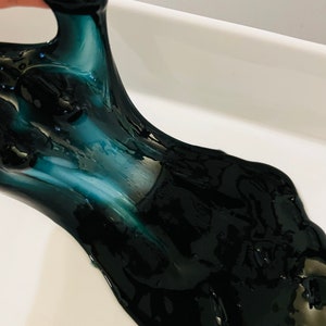 May include: Black slime with a hint of teal green, being stretched and pulled in a white ceramic dish.