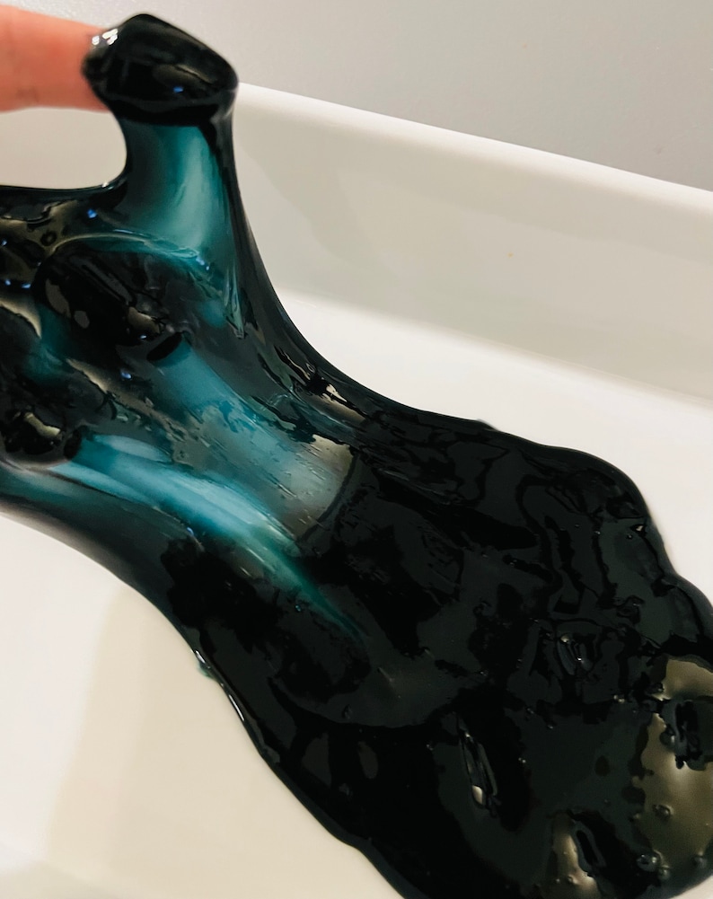 May include: Black slime with a hint of teal green, being stretched and pulled in a white ceramic dish.