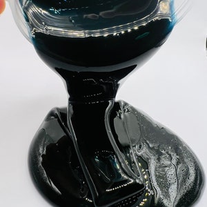 May include: Black slime being poured from a clear container. The slime is thick and gooey and has a glossy finish.