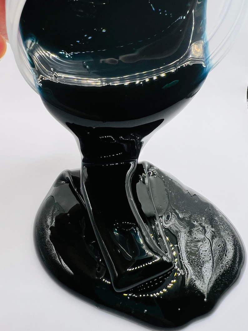 May include: Black slime being poured from a clear container. The slime is thick and gooey and has a glossy finish.