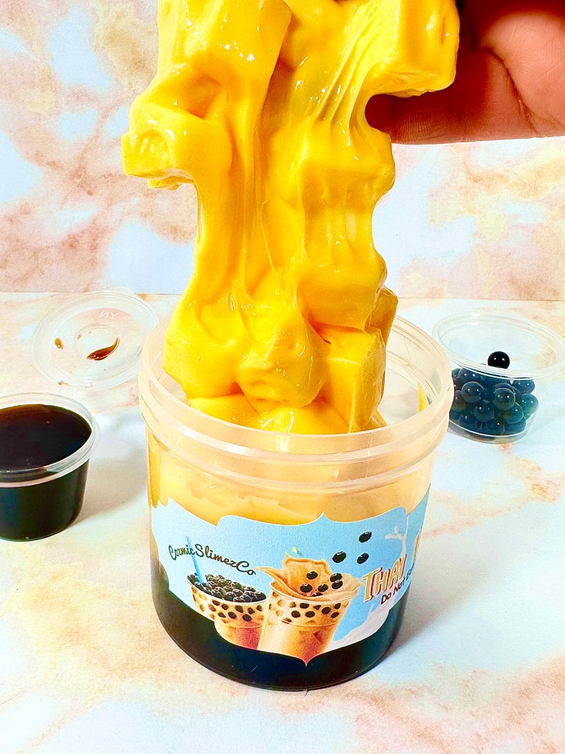 May include: A clear plastic container filled with a yellow slime that resembles a cup of bubble tea. The slime is topped with black boba pearls. The container has a label that says "Craze Slimez Co." and "Thai Tea" with a picture of two cups of bubble tea.