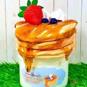 Pode incluir: Um frasco branco de argila DIY em forma de pilha de panquecas de soro de leite com um morango, chantilly e mirtilos no topo. O frasco tem um rótulo azul e branco que diz "Cosmic SlimezCo Buttermilk Pancakes Thick & Fluffy" e um rótulo menor que diz "DIY Clay Do Not Eat".