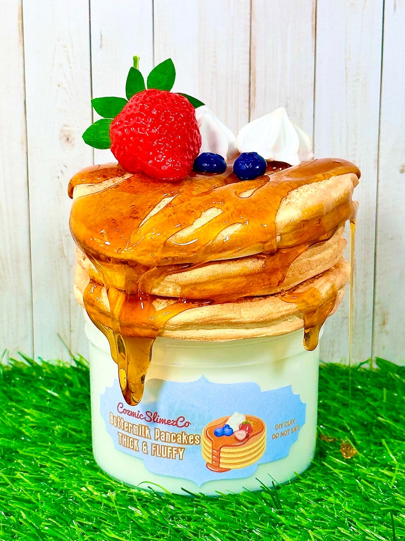 Puede incluir: Un tarro blanco de arcilla DIY con forma de pila de panqueques de suero de leche con una fresa, crema batida y ar&aacute;ndanos en la parte superior. El tarro tiene una etiqueta azul y blanca que dice "Cosmic SlimezCo Buttermilk Pancakes Thick & Fluffy" y una etiqueta m&aacute;s peque&ntilde;a que dice "DIY Clay Do Not Eat".