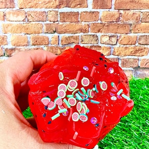 May include: Red slime with watermelon and green sprinkles. The slime is in a hand, with a brick wall and green grass in the background.