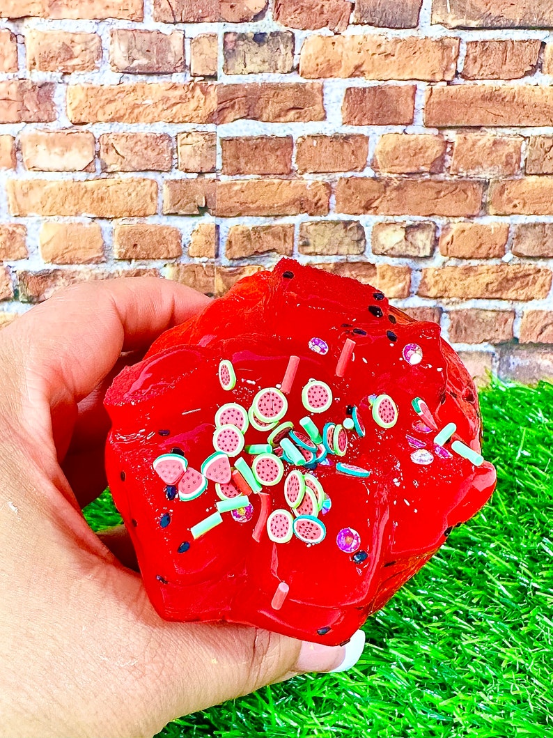 May include: Red slime with watermelon and green sprinkles. The slime is in a hand, with a brick wall and green grass in the background.