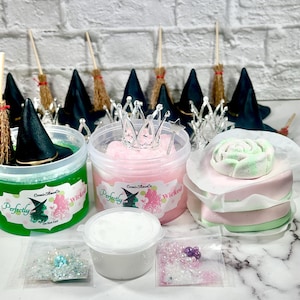 Pode incluir: Uma coleção de itens com tema de bruxa, incluindo recipientes de slime verde e rosa, chapéus de bruxa em miniatura, vassouras e uma coroa de prata. Os recipientes têm o texto "Perfectly Wicked" e um gráfico de bruxa. Também inclui um recipiente branco e sacos de glitter.