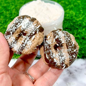 May include: Two chocolate-frosted donuts with a dark chocolate drizzle and shredded coconut topping. The donuts are held in a hand, with a jar of white powder in the background. The donuts are a light brown color.