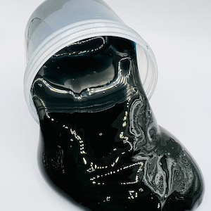May include: Black slime in a clear plastic container. The slime is spilling out of the container and onto a white surface.
