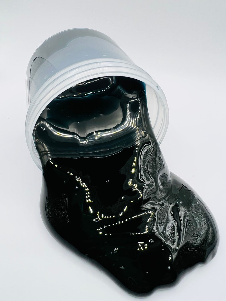 May include: Black slime in a clear plastic container. The slime is spilling out of the container and onto a white surface.