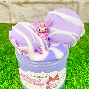 Pode incluir: Um recipiente de slime roxo decorado para se assemelhar a macarons, com listras de glacê branco e uma pequena estatueta. O recipiente tem um rótulo com o texto "Kuromi Macarons" e uma ilustração de personagem de desenho animado. As raspas adicionam cor.