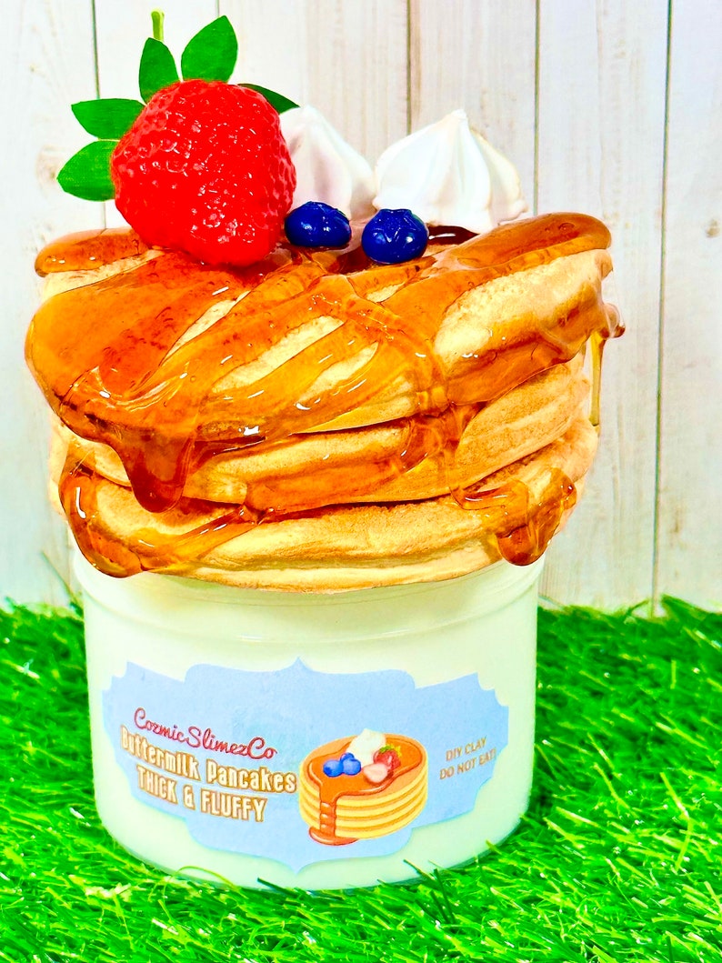 Puede incluir: Una pila de panqueques falsos de suero de leche cubiertos con jarabe de arce, ar&aacute;ndanos, crema batida y una fresa. Los panqueques est&aacute;n en una taza blanca con una etiqueta azul y blanca que dice "Cosmic SlimezCo Buttermilk Pancakes Thick & Fluffy" y "DIY Clay Do Not Eat".