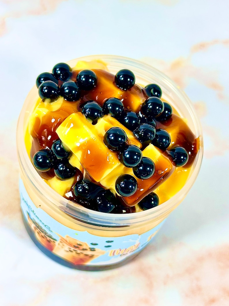 May include: A clear plastic cup filled with a yellow custard-like dessert topped with black tapioca pearls and a brown caramel sauce.