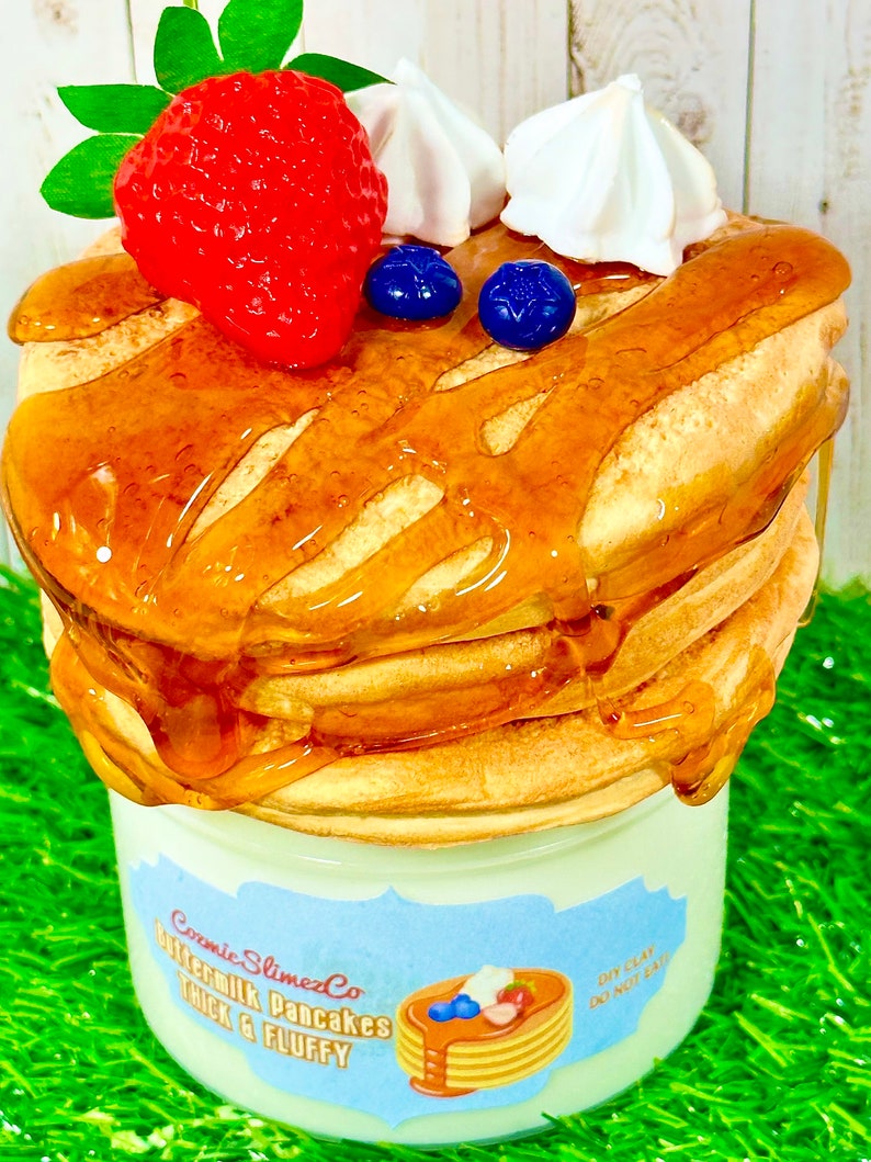 Puede incluir: Un tarro de vidrio transparente lleno de una pila de slime de panqueques de suero de leche marr&oacute;n claro cubierto con una fresa roja, merengue blanco y ar&aacute;ndanos azules. El tarro tiene una etiqueta azul que dice "Cosmic SlimezCo Buttermilk Pancakes Thick & Fluffy" y una etiqueta blanca m&aacute;s peque&ntilde;a que dice "DIY Clay Do Not Eat".
