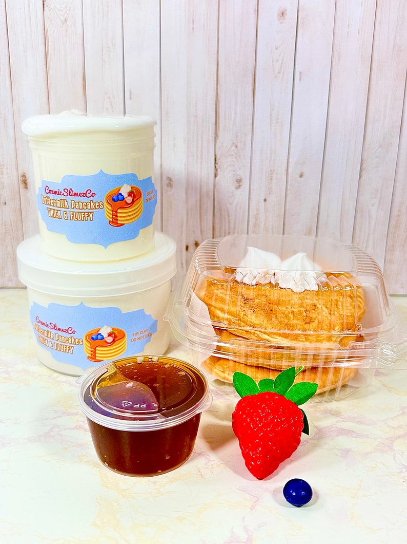 Puede incluir: Un recipiente de pl&aacute;stico blanco con una etiqueta que dice "Buttermilk Pancakes Thick & Fluffy" y una imagen de una pila de panqueques con crema batida y ar&aacute;ndanos. Hay un recipiente blanco m&aacute;s peque&ntilde;o con la misma etiqueta, un recipiente de pl&aacute;stico transparente con una pila de panqueques, un peque&ntilde;o recipiente transparente con un l&iacute;quido marr&oacute;n, una fresa roja y un ar&aacute;ndano azul.