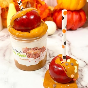 Könnte beinhalten: Zwei Karamelläpfel, einer in einem Glas und einer auf einer Oberfläche, mit dekorativen Strohhalmen. Die Äpfel sind rot mit Karamell und Streuseln. Auf dem Glas steht "SlimezCo Apple Do not eat!" Herbstliche Dekoration im Hintergrund.