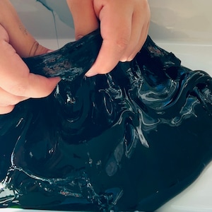 May include: A close-up of black slime being stretched and pulled by hands. The slime is shiny and has a smooth texture.