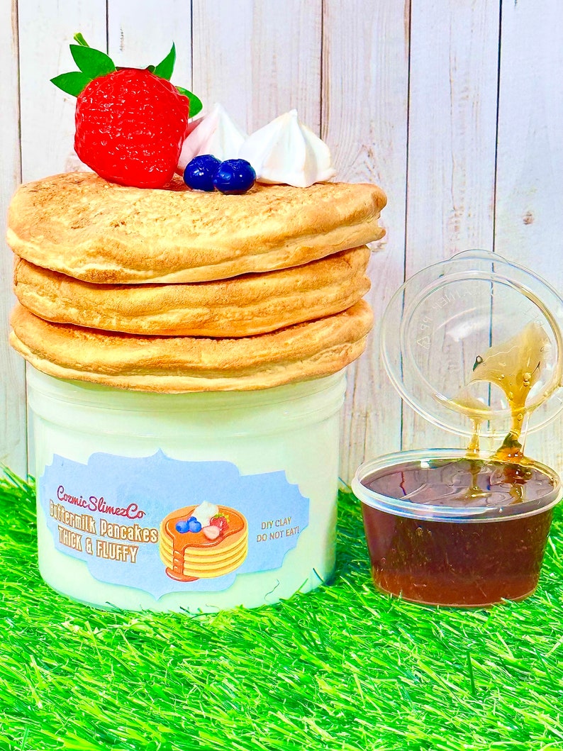 Puede incluir: Una pila de tres panqueques esponjosos de color marr&oacute;n claro, cubiertos con una fresa roja, dos ar&aacute;ndanos y dos merengues blancas. Los panqueques est&aacute;n en un recipiente blanco con una etiqueta azul que dice "Cosmic SlimezCo Buttermilk Pancakes Thick & Fluffy" y una etiqueta m&aacute;s peque&ntilde;a que dice "DIY Clay Do Not Eat". Un recipiente de pl&aacute;stico transparente con un l&iacute;quido marr&oacute;n, probablemente jarabe, se est&aacute; vertiendo sobre los panqueques.