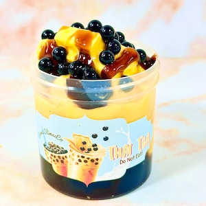 May include: A clear plastic container filled with a yellow slime topped with black boba pearls and a brown chocolate sauce. The container has a label that reads "Thai Tea Do Not Eat".