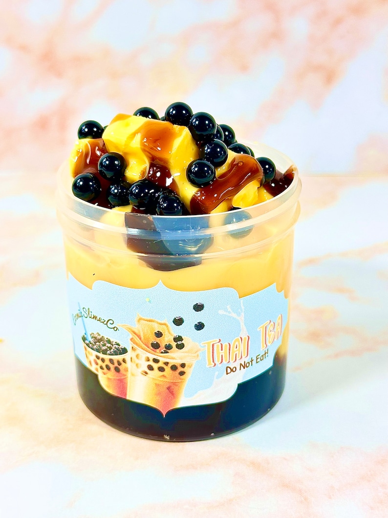 May include: A clear plastic container filled with a yellow slime topped with black boba pearls and a brown chocolate sauce. The container has a label that reads "Thai Tea Do Not Eat".