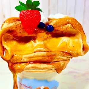 Puede incluir: Primer plano de un tarro de cer&aacute;mica blanco con una etiqueta azul y blanca que dice "Cosmic Slumber Co. Buttermilk Pancakes Thick & Fluffy". El tarro est&aacute; lleno de una masa de panqueques amarilla y esponjosa cubierta con una fresa roja, ar&aacute;ndanos y un chorrito de jarabe de arce.