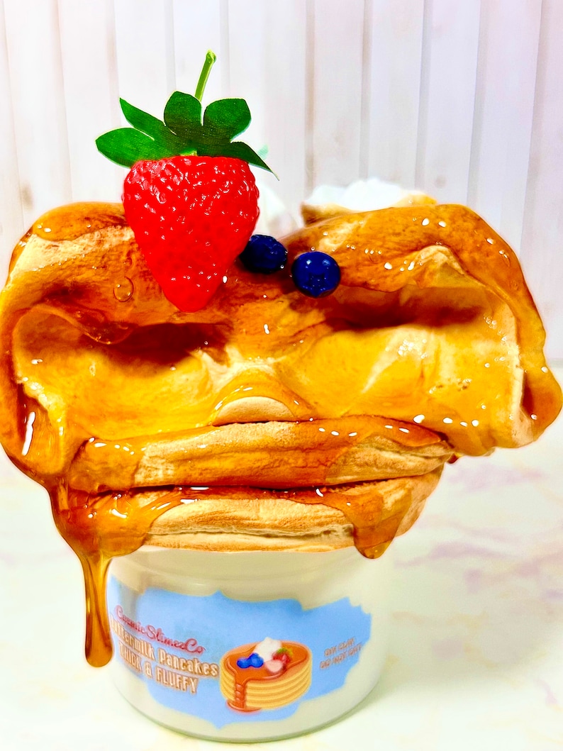 Puede incluir: Primer plano de un tarro de cer&aacute;mica blanco con una etiqueta azul y blanca que dice "Cosmic Slumber Co. Buttermilk Pancakes Thick & Fluffy". El tarro est&aacute; lleno de una masa de panqueques amarilla y esponjosa cubierta con una fresa roja, ar&aacute;ndanos y un chorrito de jarabe de arce.