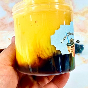 May include: A clear plastic jar filled with a yellow mango-flavored slime with black tapioca pearls at the bottom. The jar has a blue and white label that says "Cosmic Slime Co."