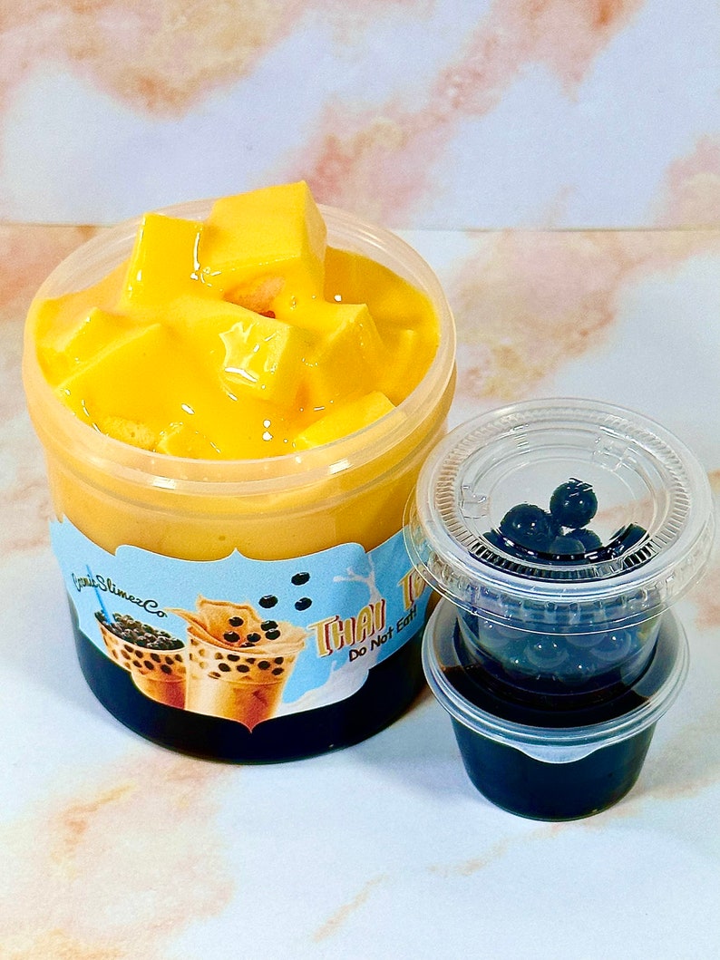 May include: A clear plastic container filled with yellow mango jelly and topped with a layer of mango pudding. A smaller clear plastic container with a black lid holds black tapioca pearls. The label on the larger container reads "Thai Tea Do Not Eat".