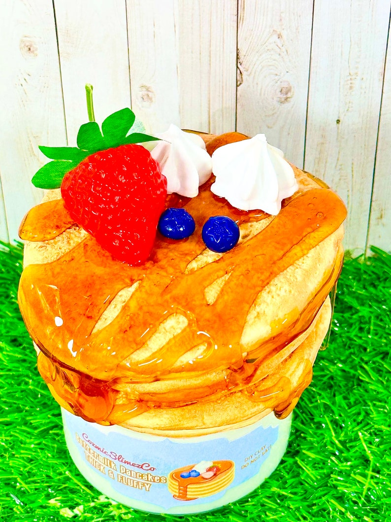 Puede incluir: Primer plano de un tarro de slime con aroma a panqueque con una pila de panqueques, crema batida, una fresa y ar&aacute;ndanos en la parte superior. El tarro est&aacute; etiquetado como "Cosmic Slime Co. Buttermilk Pancakes Topped & Fluffy".