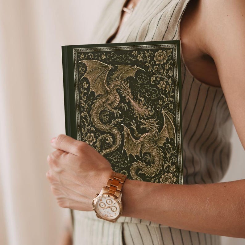 May include: A hardback book with a dark green cover featuring an intricate gold-toned illustration of two dragons facing each other amidst floral patterns and clouds. The design includes detailed scales, wings, and fire breath in a metallic finish.