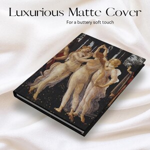 May include: A rectangular hardcover notebook featuring a matte finish with a reproduction of Botticelli's Three Graces painting on the front cover. The text "Luxurious Matte Cover" and "For a buttery soft touch" is printed above the notebook.