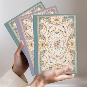 May include: Three rectangular hardcover journals featuring a decorative cream-colored center panel with intricate gold and brown scrollwork, floral motifs, and a border in shades of blue and lavender, bound with solid-colored spines in blue and purple.