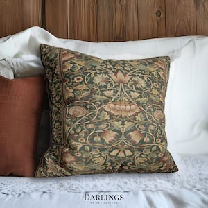 May include: A square cushion with a detailed floral pattern in green, brown, and beige tones. The cushion is placed on a white bed, showcasing a botanical design. The brand name "DARLINGS OF ART DESIGNS" is visible at the bottom of the image.