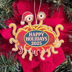 Op de afbeelding: Een grillige kerstversiering. De ornament toont een cartoonachtig wezen met gele tentakels, wiebelogen en een kerstmuts. Het heeft een rode body en een blauwe ovaal met de tekst "HAPPY HOLIDAYS 2025".